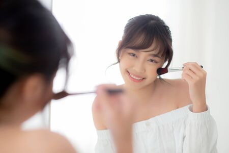 Beauty portrait young asian woman smile with face looking mirror applying makeup with brush cheek in the bedroom, beautiful of girl holding blusher, skin care and cosmetic concept.の写真素材