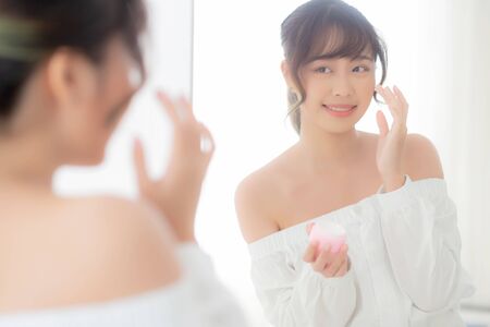 Beautiful portrait young asian woman applying cream moisturizer or lotion skin care cosmetic on face looking mirror, girl with treatment facial, health and wellness concept.の写真素材