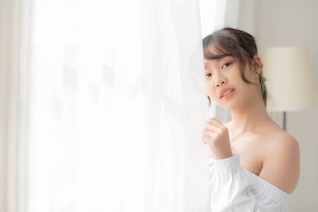 Portrait of beautiful young asian woman standing the window and smile while wake up with sunrise at morning, girl happy with freshness and cheerful, lifestyle and relax concept.の写真素材