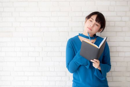Beautiful asian woman smiling standing thinking and writing notebook on concrete cement white background at home, girl homework on book, education and lifestyle concept.の写真素材