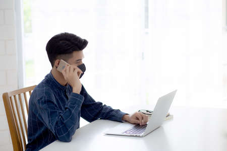 Young asian business man in face mask talking smartphone and work on laptop computer for protect covid-19, businessman quarantine work from home with notebook, social distancing and stay home.の写真素材