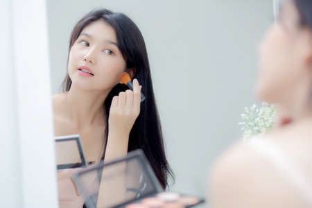 Beauty young asian woman happy and smile with face looking mirror applying makeup with brusher of cheek in the room, beautiful of girl holding brush make-up on facial, skin care and cosmetic concept.の写真素材