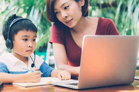 Young asian mother and son using laptop computer for study and learning together at home, boy writing on notebook for homework and wearing headphone, teacher or mom support child, education concept.の写真素材
