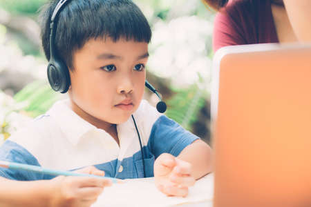 Young asian mother and son using laptop computer for study and learning together at home, boy writing on notebook for homework and wearing headphone, teacher or mom support child, education concept.の写真素材