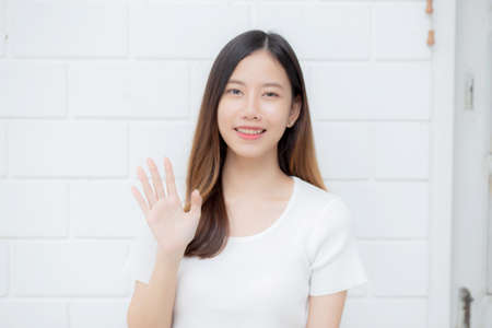 Beautiful young asian woman confident gesture say hello or greeting with waving her hand on cement background at home, friendly welcome, portrait girl cheerful expression happy and satisfied.の写真素材