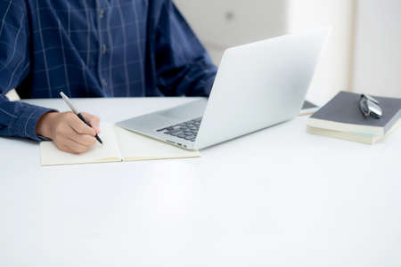 Closeup hand of business man writing on note while using laptop computer on desk at home, male...