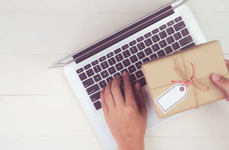 Hand of man holding gift box and tag having laptop computer on wooden table, present giving for...