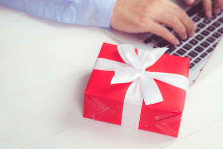 Hand of man holding red gift box and having laptop computer on wooden table, online shopping for...