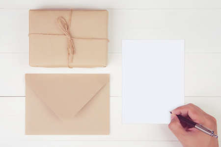 Hand of man writing letter with blank greeting card and having gift box with kraft paper on wooden...