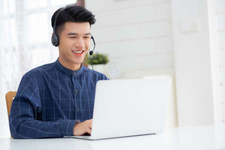 Young asian businessman working on laptop computer wearing headphone at home, business man wearing headset for video conference, communication and education, male study and learning for e-learning.の写真素材
