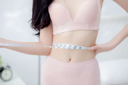 Beautiful young asian woman sexy body slim measuring abdomen for control weight loss in the room, beauty girl belly thin have cellulite with tape measure for diet, health and wellness concept.の写真素材