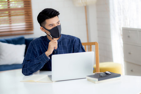 Young business man in face mask planning and working from home on laptop computer on desk for protection for outbreak of covid-19, male quarantine stay home using notebook on desk, new normal.の写真素材