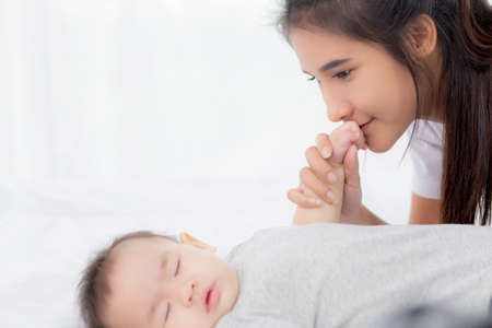 Young asian mother kiss hand of little baby girl with tender on bed in the bedroom, mom love newborn...