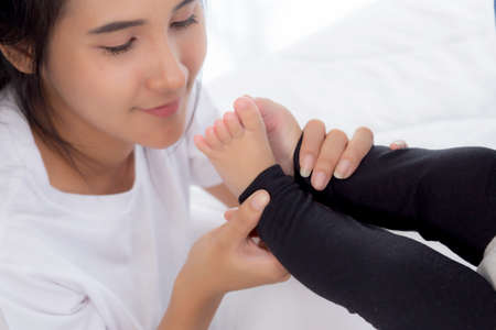 Young asian mother kiss foot of little baby girl on bed in bedroom at home, mom and newborn with...