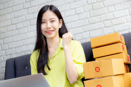 Young asian woman is merchant using laptop computer on sofa checking order of customer with purchase online shopping, business SME or startup, entrepreneur or seller and service delivery product.の写真素材