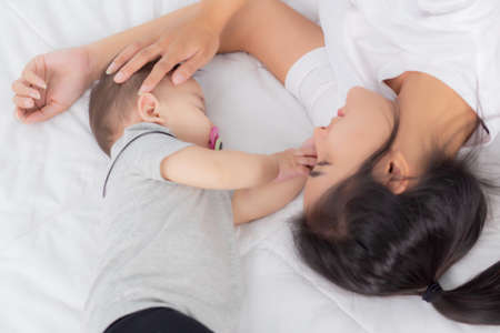 Young asian mother touch head of little baby girl with tender on bed in the bedroom, mom love newborn and care, woman with expression with child together, parent and daughter, family concept.の写真素材