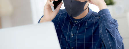 Young asian business man in face mask talking smartphone and work on laptop computer for protect...