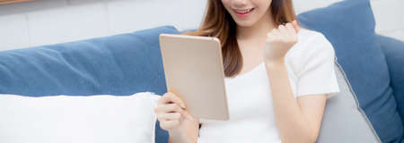 Young attractive asian woman resting using browsing tablet computer on sofa at home, happy girl sitting on couch relax reading digital gadget with excited at house, communication and lifestyle concept.の写真素材