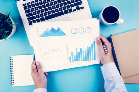 Hand of businessman holding documents report statistic financial with graph and chart and laptop computer and coffee on deskの写真素材