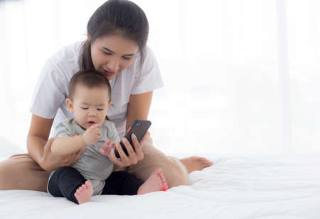 Young asian mother and little baby girl or newborn selfie with smart phone on bed in bedroom