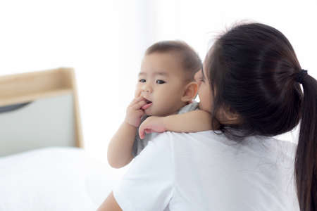 Young asian mother holding little baby girl together in the bedroom at home, mom carry child and care, woman and kid with carefree, toddler and parent, emotion and expression, family concept.の写真素材