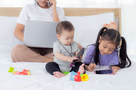 Young asian mother working at home online to internet with laptop computer and talking phone and daughter looking tablet on bed at bedroom, business woman is freelance, new normal, family concept.の写真素材