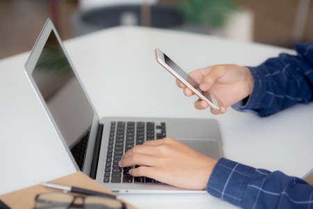 Closeup hand of young man working laptop computer and reading smartphone on internet online on desk...