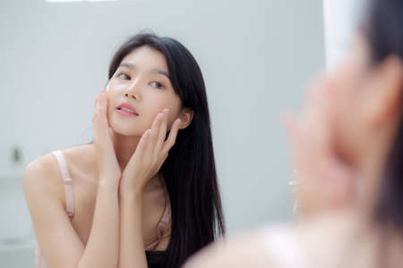 Beautiful young asian woman smiling look at mirror of checking face with skin care and cosmetic for rejuvenation and hygiene, beauty girl happy clean facial with cream or lotion and for health.の写真素材