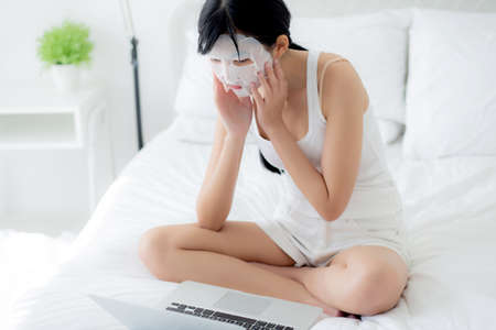 Beautiful young asian woman with sheet facial mask sitting working on laptop computer on bed at bedroom, beauty girl applying face mask for skincare of wrinkle at home, skin care, health and wellness.の写真素材