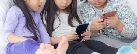Happy asian little kid and friends playing smartphone on sofa at home, children using phone together on couch, girl watching smart phone for entertainment, lifestyle and communication concept.の写真素材