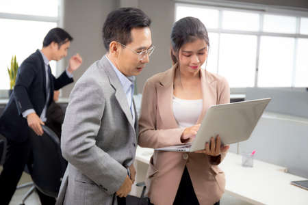 Asian businessman and businesswoman talking and looking laptop computer in the office, man standing explaining about financial with woman together, entrepreneur and employee planning about business.の写真素材