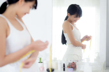 Beautiful young asian woman standing and measuring tape with waist for control weightloss looking mirror in the room, beauty asia girl figure thin with dieting, weight loss and healthy concepts.の写真素材