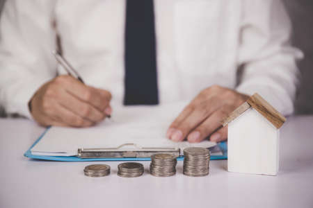 Hands of business man sign agreement buying home while model home and stack of coins on desk, finance for residential and success, loan and mortgage, investment and insurance for real estate.の写真素材