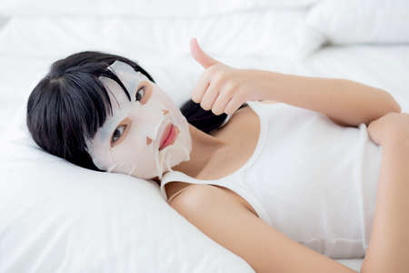 Beautiful young asian woman with sheet facial mask and thumbs up on bed at bedroom, beauty girl lying applying cosmetic and makeup with skin care face for wrinkle, skincare treatment and health.の写真素材