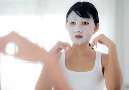 Beautiful young asian woman with sheet facial mask and looking mirror at home, beauty girl applying cosmetic and makeup with skin care face for wrinkle, treatment and moisturizer, health and wellness.の写真素材