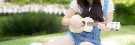 Happiness young asian woman sitting and playing ukulele while relaxation for entertainment is hobby on summer in the park, girl is musician doing activity with feeling positive, lifestyles concept.の写真素材