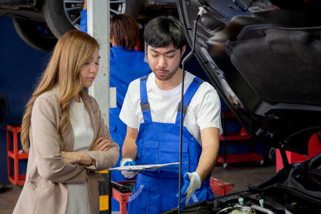 Young mechanic talking to customer asian woman about car with repairing in garage, auto service, technician man explaining problem of vehicle and look clipboard with client, transportation concept.の写真素材