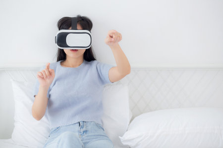 Young asian woman wearing vr headset playing game with excited on bed in the bedroom at home, female...