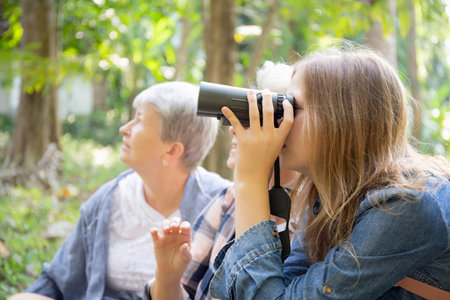Teenager girl and two grandmother traveling travel to forest and adventure while using binoculars exploration together with happy, senior woman and granddaughter hiking with fun and vitality.の写真素材