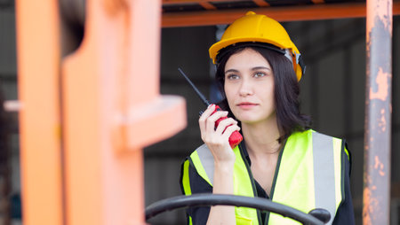 Young asian woman is foreman using radio for communication while driving forklift at distribution warehouse, engineer working and shipping container at factory, cargo and logistic, industry concept.の写真素材