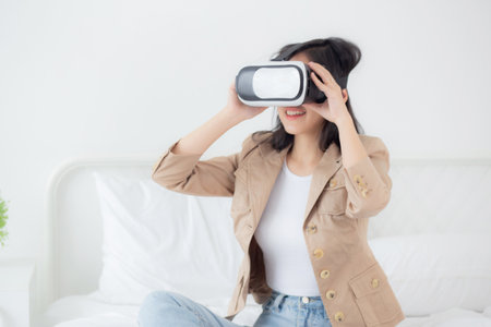 Young asian woman wearing vr headset playing game with excited on bed in the bedroom at home, female using virtual reality or metaverse innovation for simulation 3D, lifestyles and technology concept.の写真素材