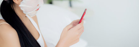 Beautiful young asian woman with sheet facial mask sitting watching smartphone on bed at bedroom, beauty girl applying face mask for skincare of wrinkle at home, skin care, health and wellness.の写真素材