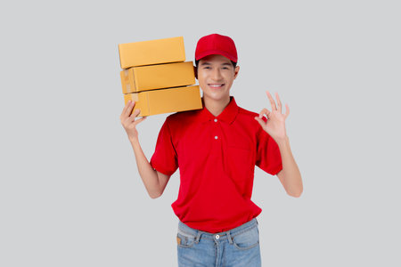 Young asian man in uniform red and cap standing carrying box stack and gesture ok isolated white background, employee hold cargo, courier and delivery, transportation and service, logistic and cargo.の写真素材