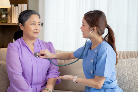 Young asian nurse or caregiver sitting on sofa holding stethoscope listen heartbeat with senior woman in living room at home, doctor check cardiologist disease with elderly, medical and insurance.の写真素材