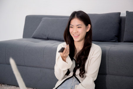 Beautiful young asian woman sitting using remote control with television for watching series with...