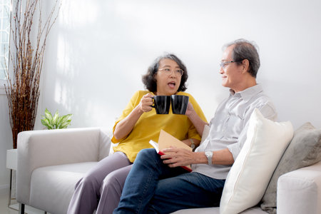 Happiness senior couple sitting on sofa cheer coffee cup and drinking in living room at home, happy elderly man and woman sitting on couch drinking coffee and talking with cozy, bonding and relation.の写真素材