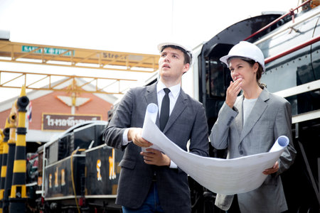 Young caucasian engineer man and woman in suit checking train looking blueprint in station, team...
