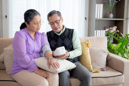 Asian senior couple sitting on sofa checking blood pressure with pressure gauge in living room at home, elderly man and woman sitting on couch checkup health and pressure, medical of patient.の写真素材