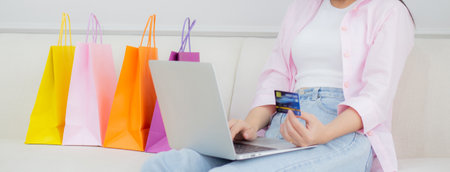 Young asian woman sitting on sofa using laptop computer shopping online with credit card while paper bag on sofa, female paying with transaction financial, purchase and payment, business concept.の写真素材