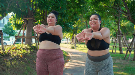 Obese two young asian woman doing stretching arm muscle in the park, woman with overweight warm up and exercise for dieting and health, workout and motivation, oversized and challenge, plus size.の写真素材